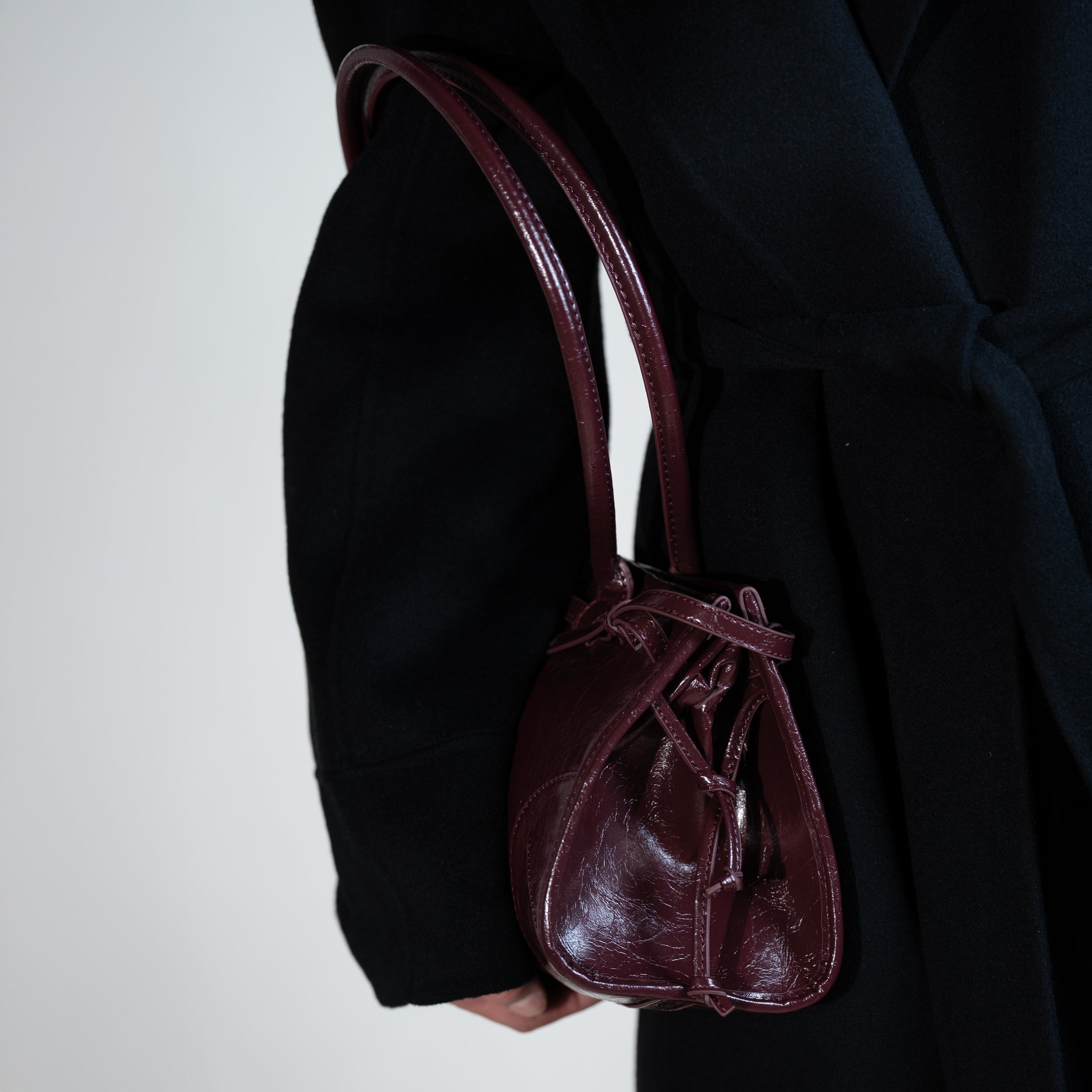 sac à main forme baguette femme en cuir verni bordeaux vintage porté à la main sur un manteau noir 