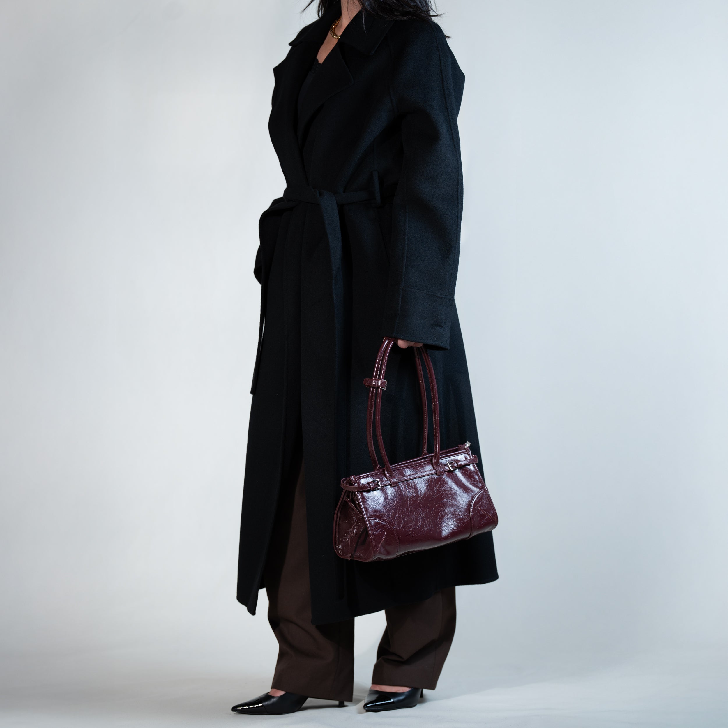 sac à main forme baguette femme en cuir verni bordeaux vintage porté à la main sur un manteau noir, un pantalon de tailleur marron et des escarpins vernis