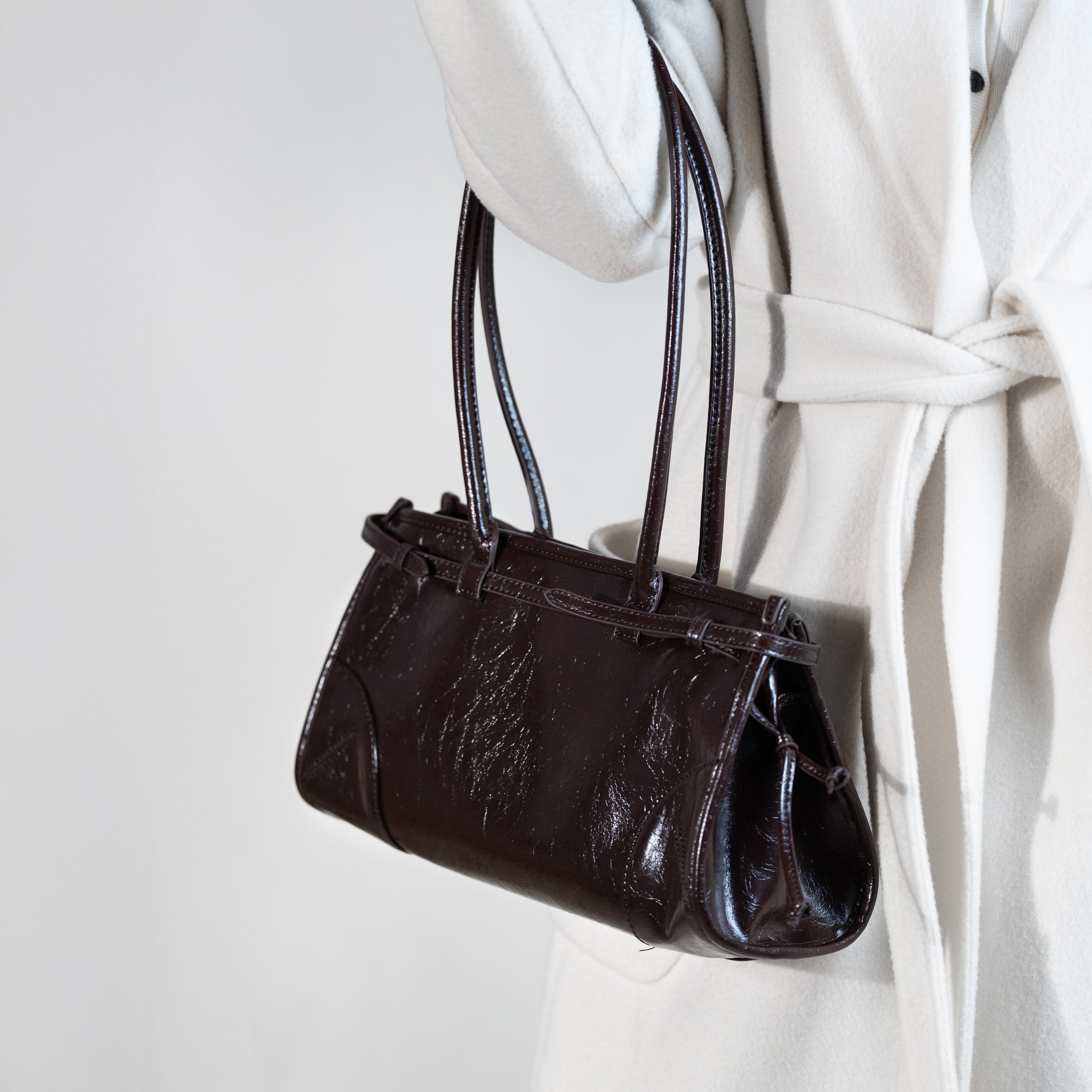 sac à main forme baguette femme en cuir verni marron porté au creux de bras sur un manteau blanc