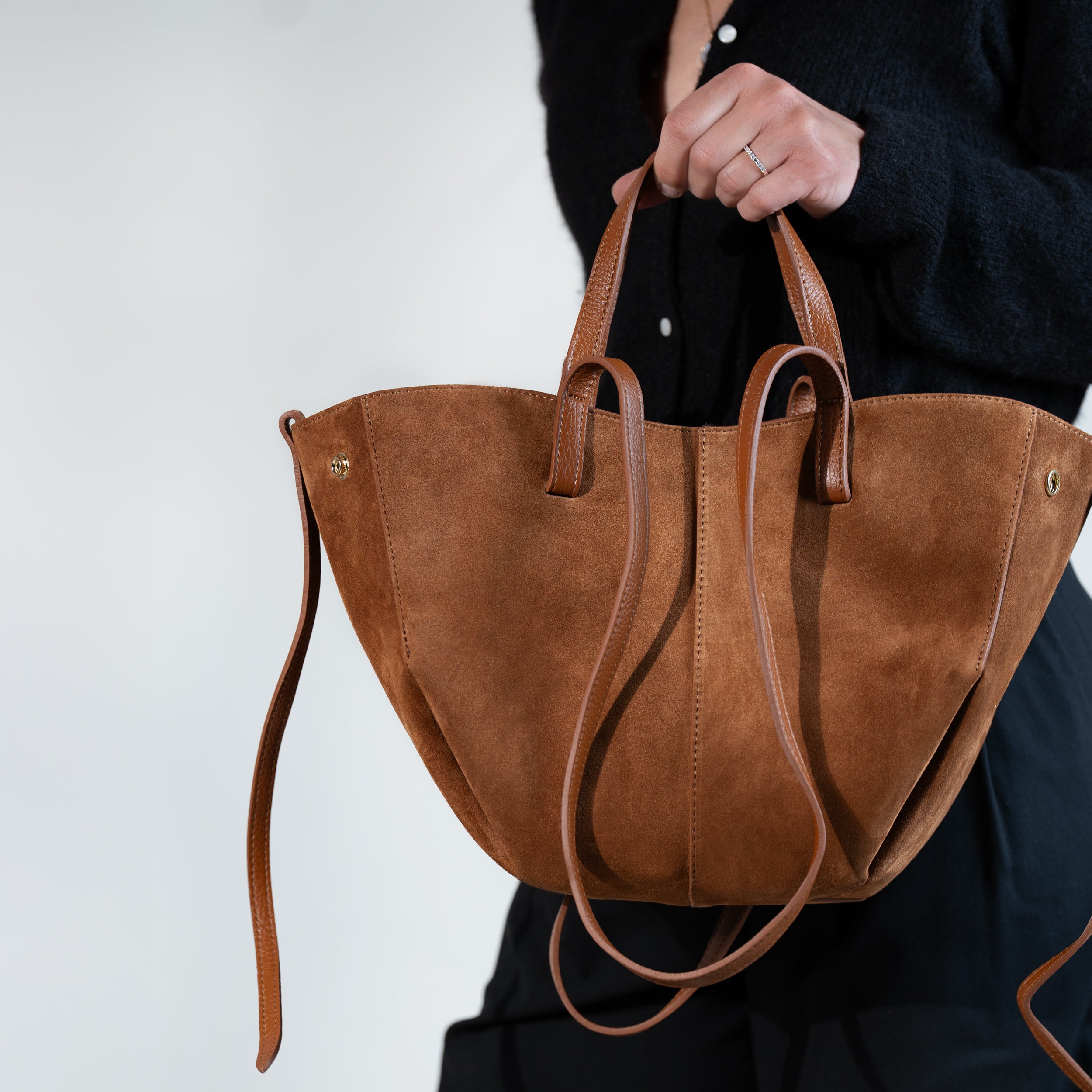 sac cabas forme trapèze en cuir suédé ou daim camel porté main sur un pull noir et un pantalon noir