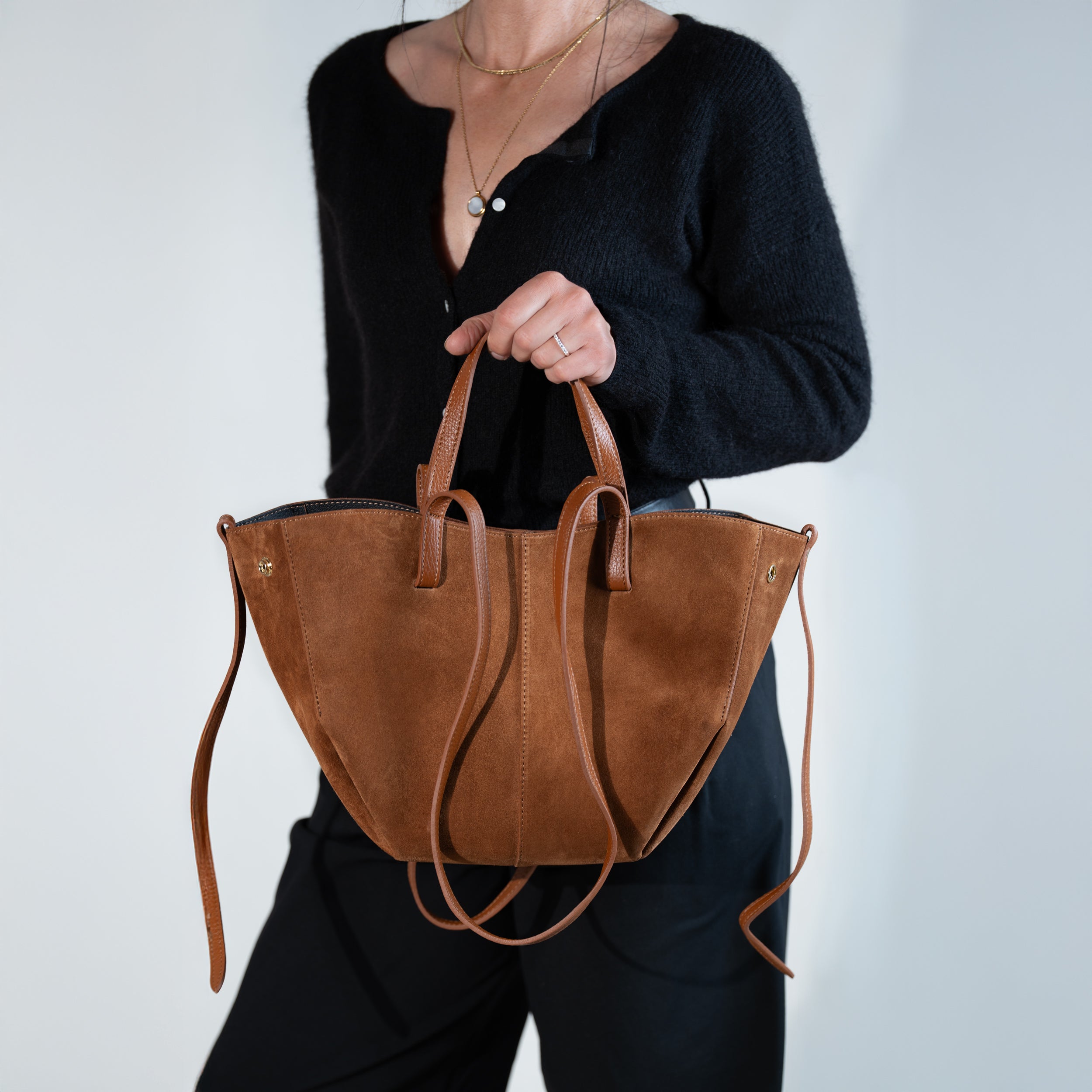 sac cabas pour femme forme trapèze en cuir suédé ou daim camel porté main sur un pull noir et un pantalon noir