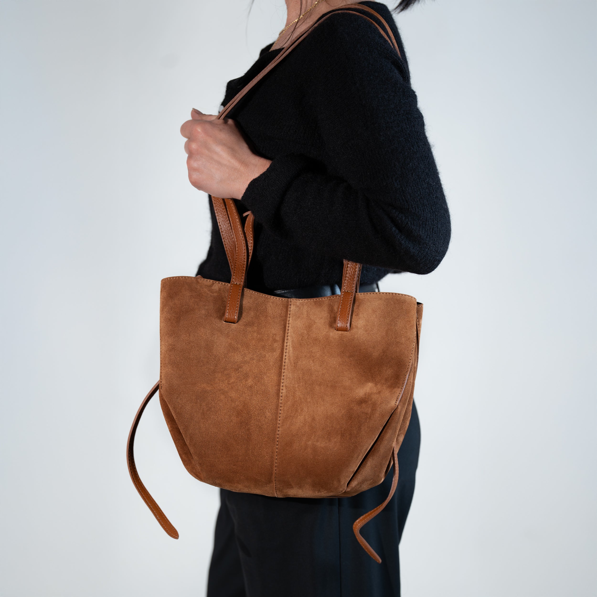 sac cabas pour femme forme trapèze en cuir suédé ou daim camel porté main sur un pull noir et un pantalon noir 