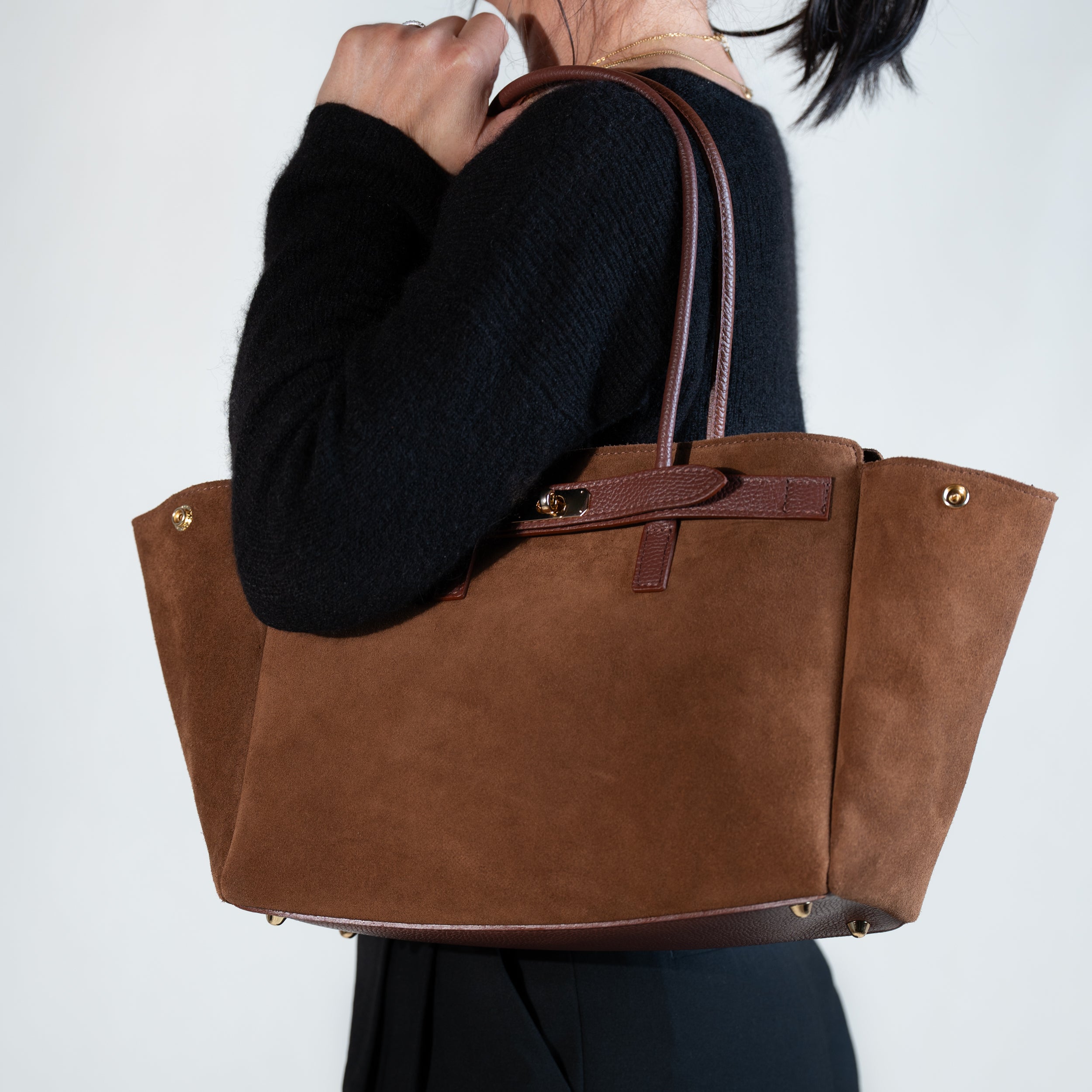grand sac cabas femme en cuir suédé ou daim camel en forme trapèze avec fermoir tourniquet porté à l'épaule sur une tenue avec un pull noir et un pantalon tailleur noir