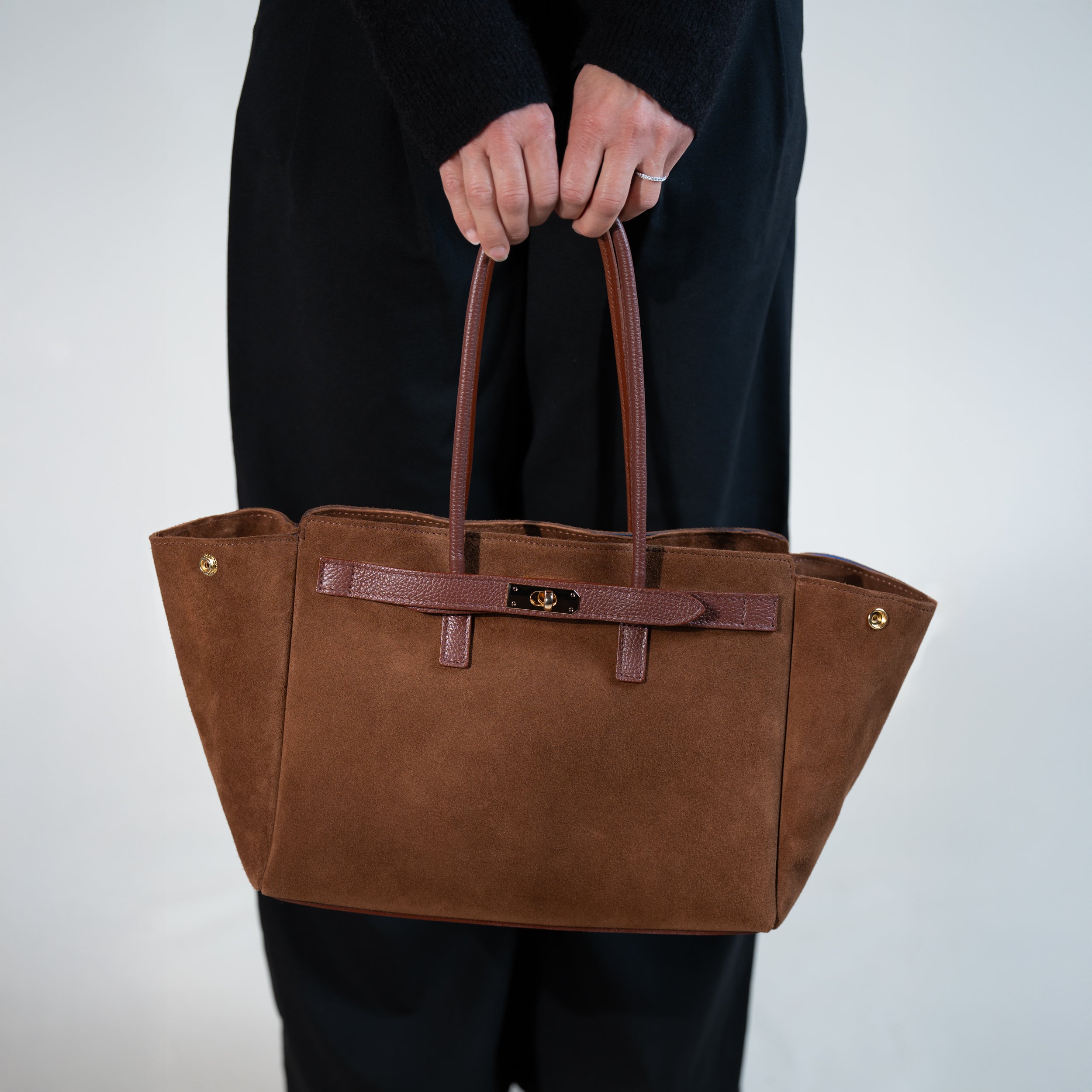 grand sac cabas femme en cuir suédé ou daim camel en forme trapèze avec fermoir tourniquet porté à la main sur un pantalon tailleur noir 