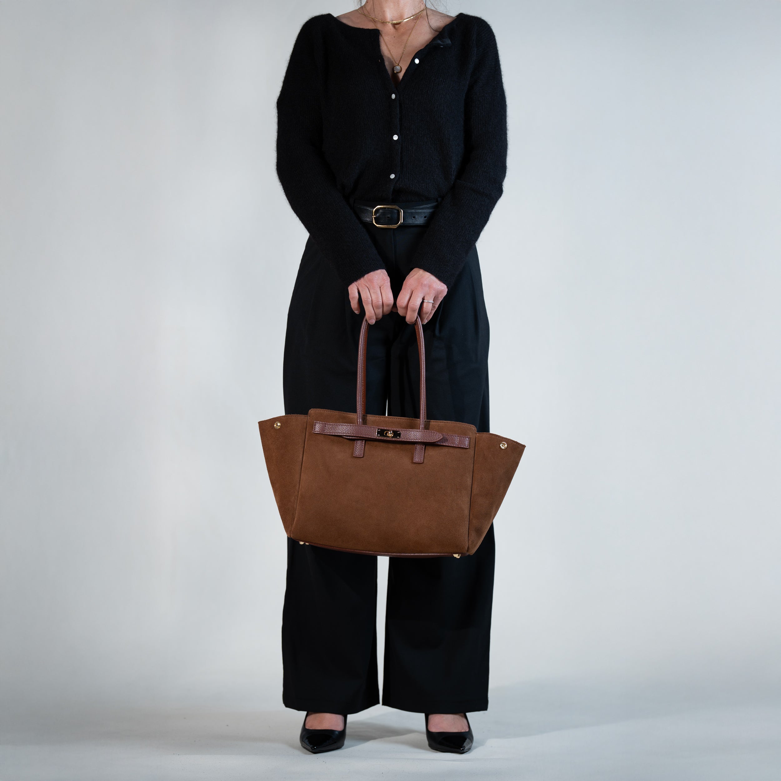 sac cabas femme en cuir suédé ou daim camel en forme trapèze avec fermoir tourniquet porté à la main sur une tenue avec un pull noir, une ceinture noir, des escarpins vernis et un pantalon tailleur noir
