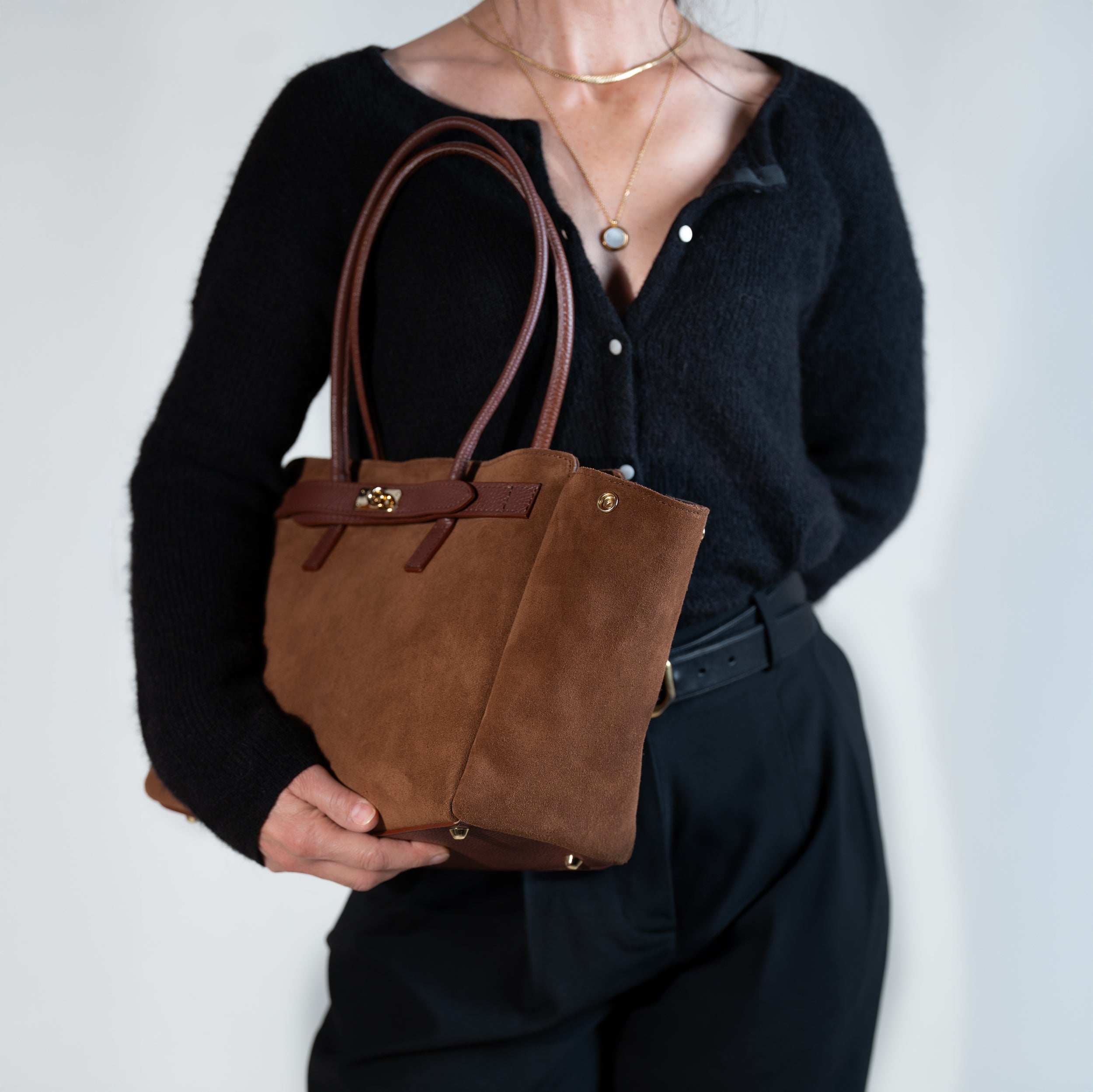 sac cabas femme en cuir suédé ou daim camel en forme trapèze avec fermoir tourniquet et clous de fond de sac, porté au bras sur une tenue avec un pull noir, une ceinture noir et un pantalon tailleur noir