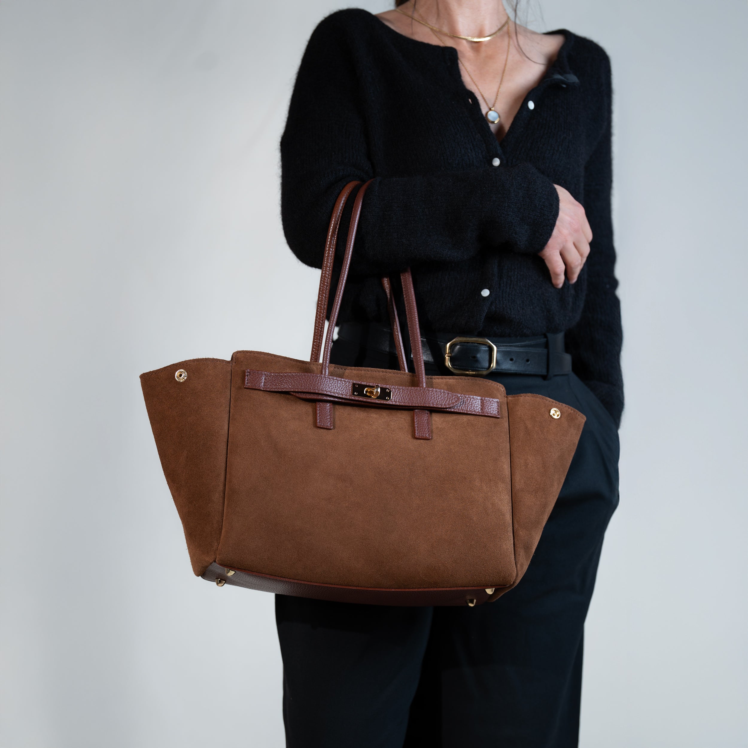 sac cabas femme en cuir suédé ou daim camel en forme trapèze avec fermoir tourniquet porté au bras sur une tenue avec un pull noir, une ceinture noir et un pantalon tailleur noir