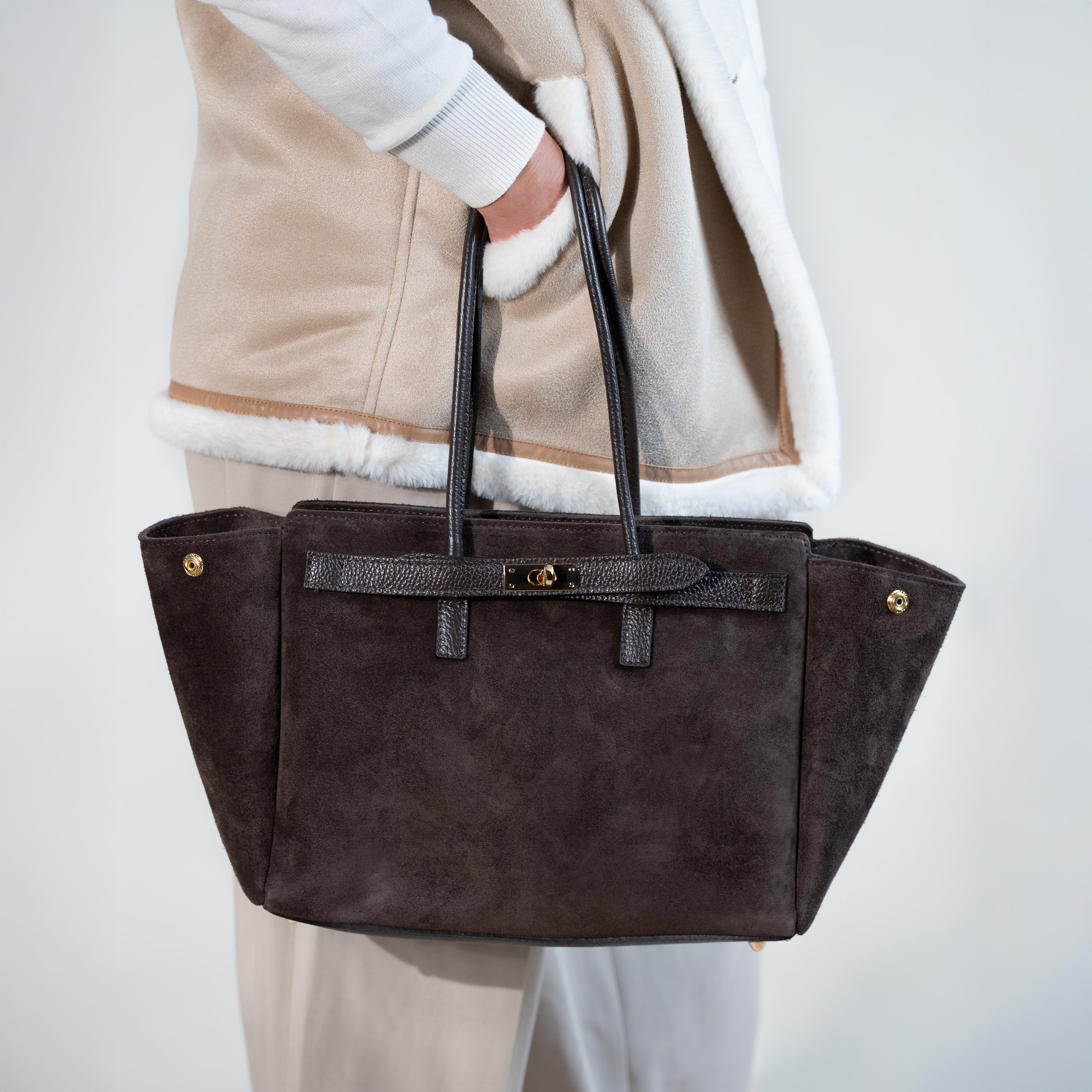 sac cabas femme en cuir suédé ou daim brun en forme de trapèze avec fermoir tourniquet porté à la main sur un pantalon tailleur beige et un manteau en daim beige