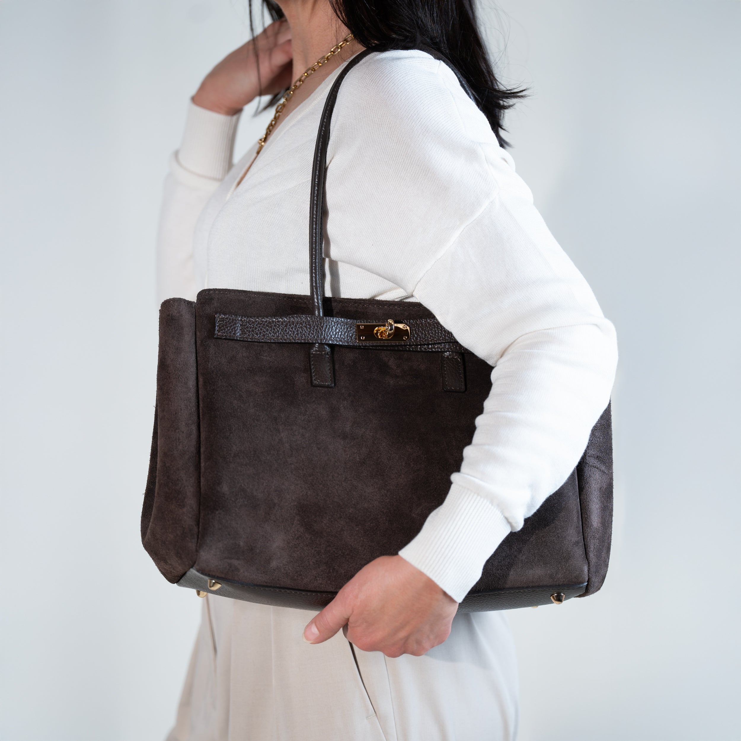 sac cabas femme en cuir suédé ou daim brun en forme rectangulaire avec fermoir tourniquet porté à l'épaule sur un pull blanc et un pantalon beige
