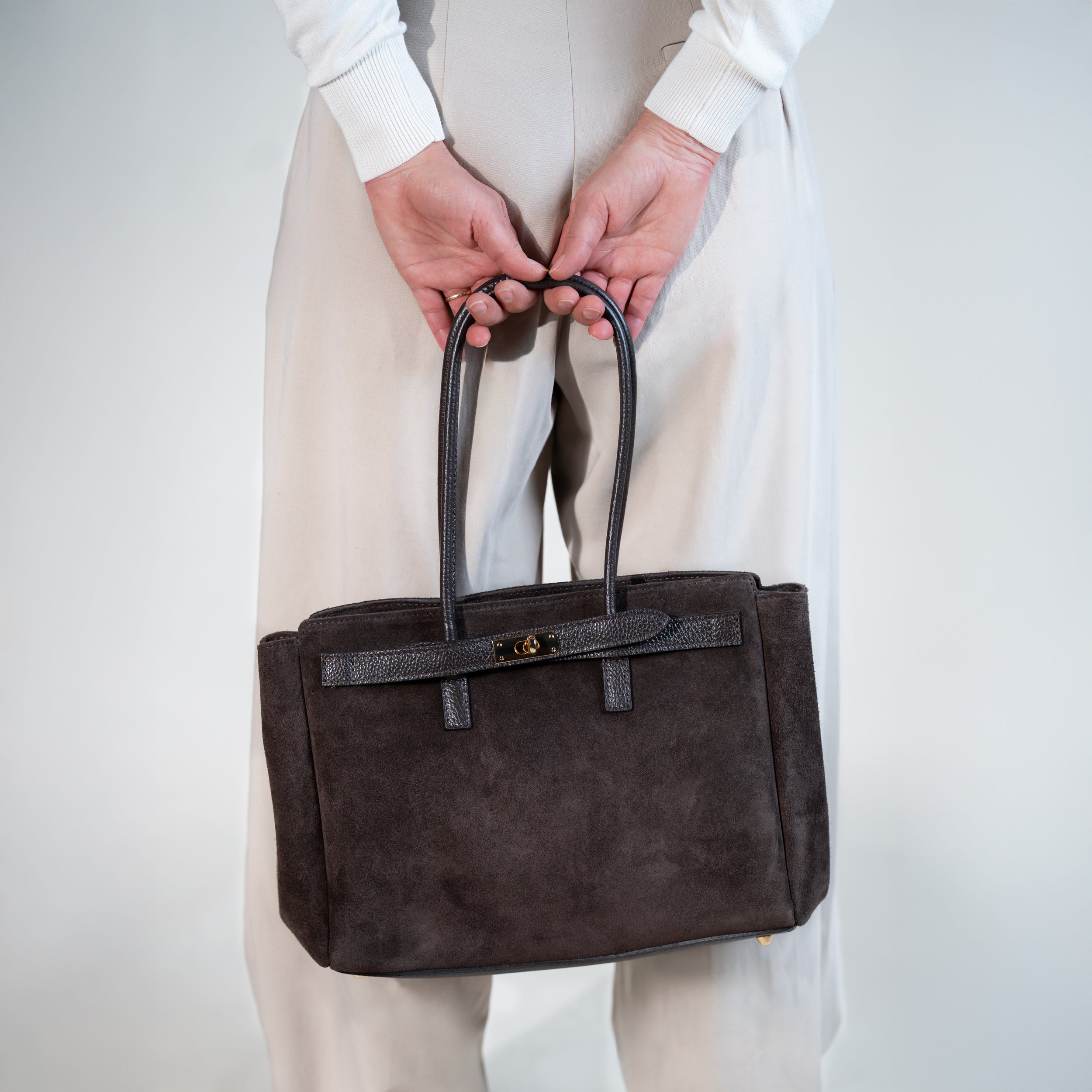 sac cabas femme en cuir suédé ou daim brun en forme rectangulaire avec fermoir tourniquet porté à la main sur un pantalon tailleur beige