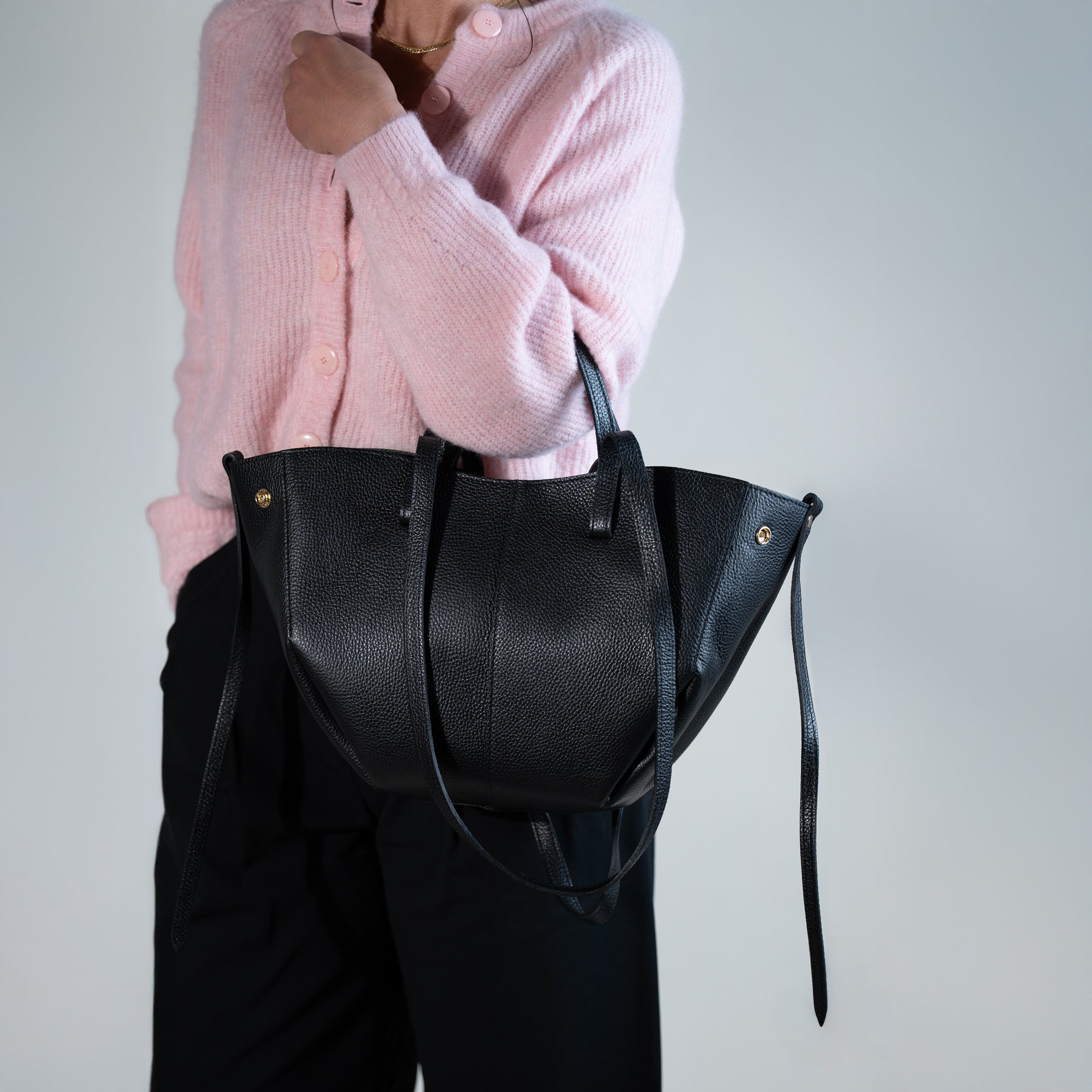 grand sac cabas pour femme forme trapèze en cuir lisse noir porté au creux de bras avec pull rose et pantalon noir