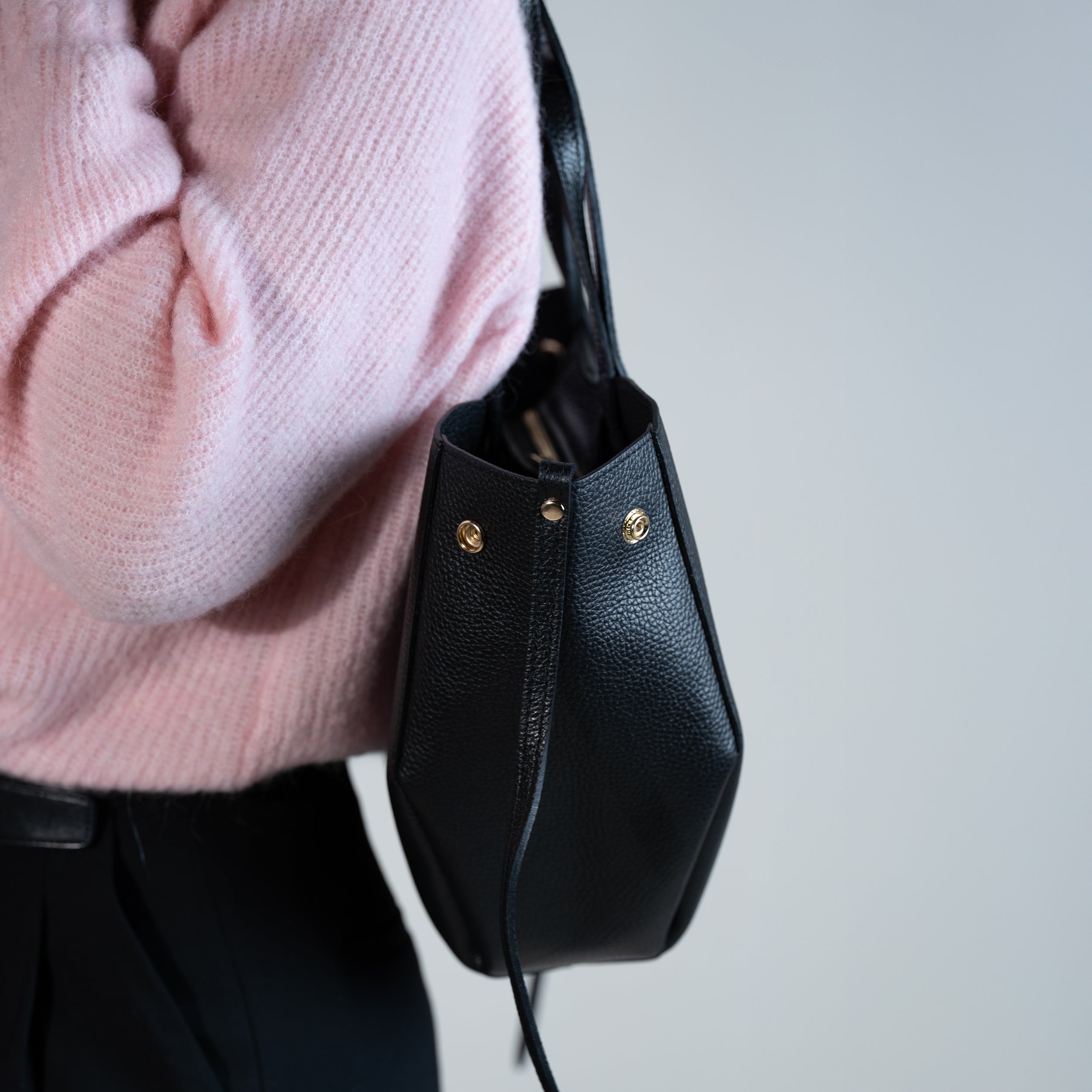côté d'un sac cabas pour femme forme trapèze en cuir lisse noir porté à l'épaule avec pull rose et pantalon noir