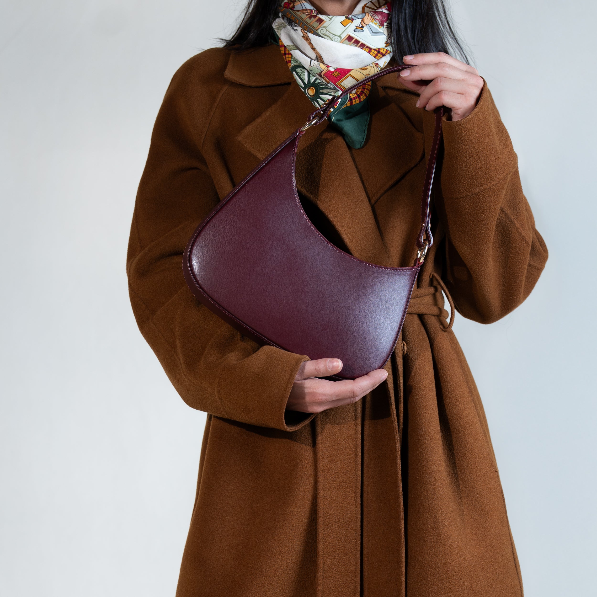 sac à main forme hobo demi lune asymétrique pour femme en cuir lisse bordeaux porté à la main sur manteau camel et foulard en soie