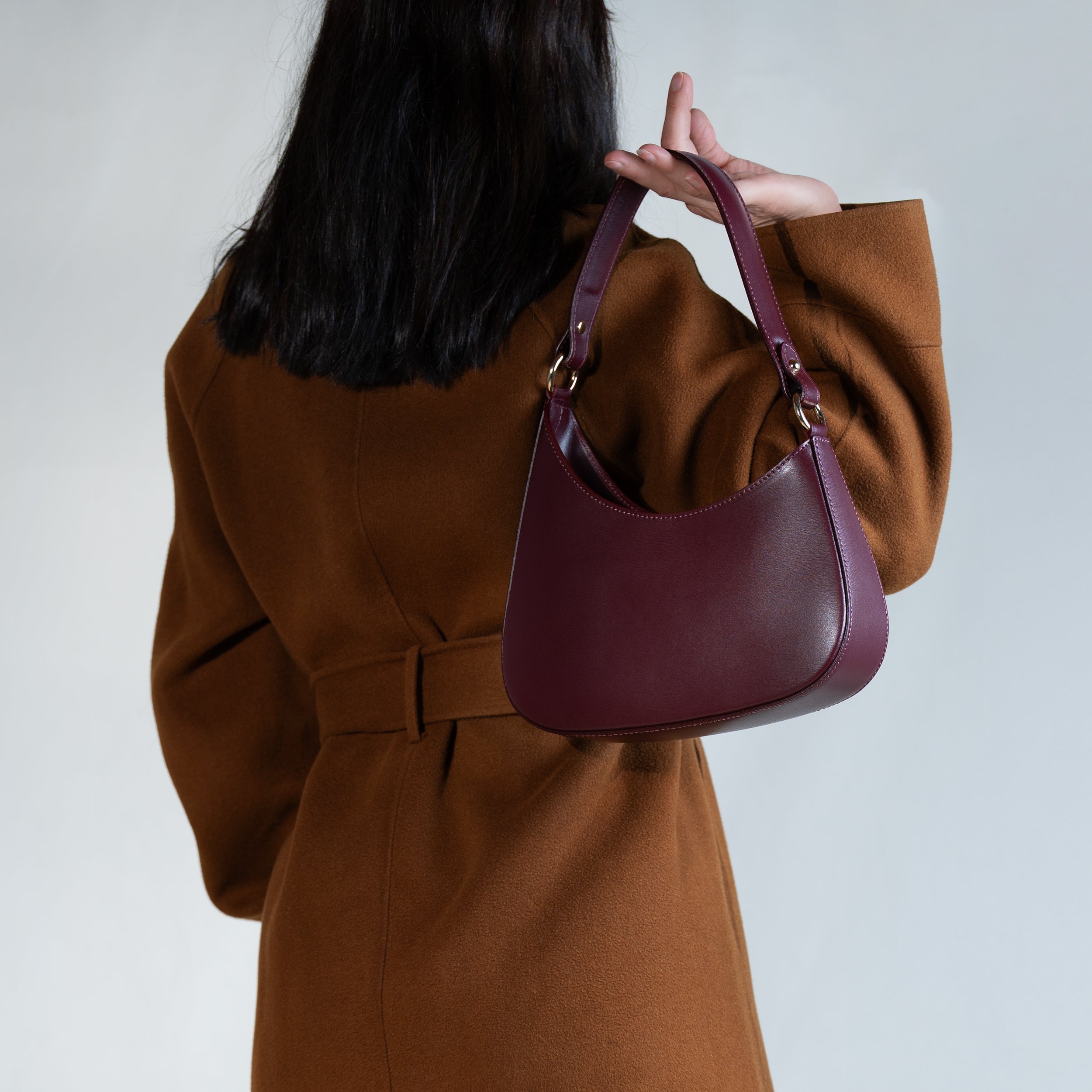 sac à main forme hobo demi lune asymétrique pour femme en cuir lisse bordeaux porté à la main de dos sur manteau camel 