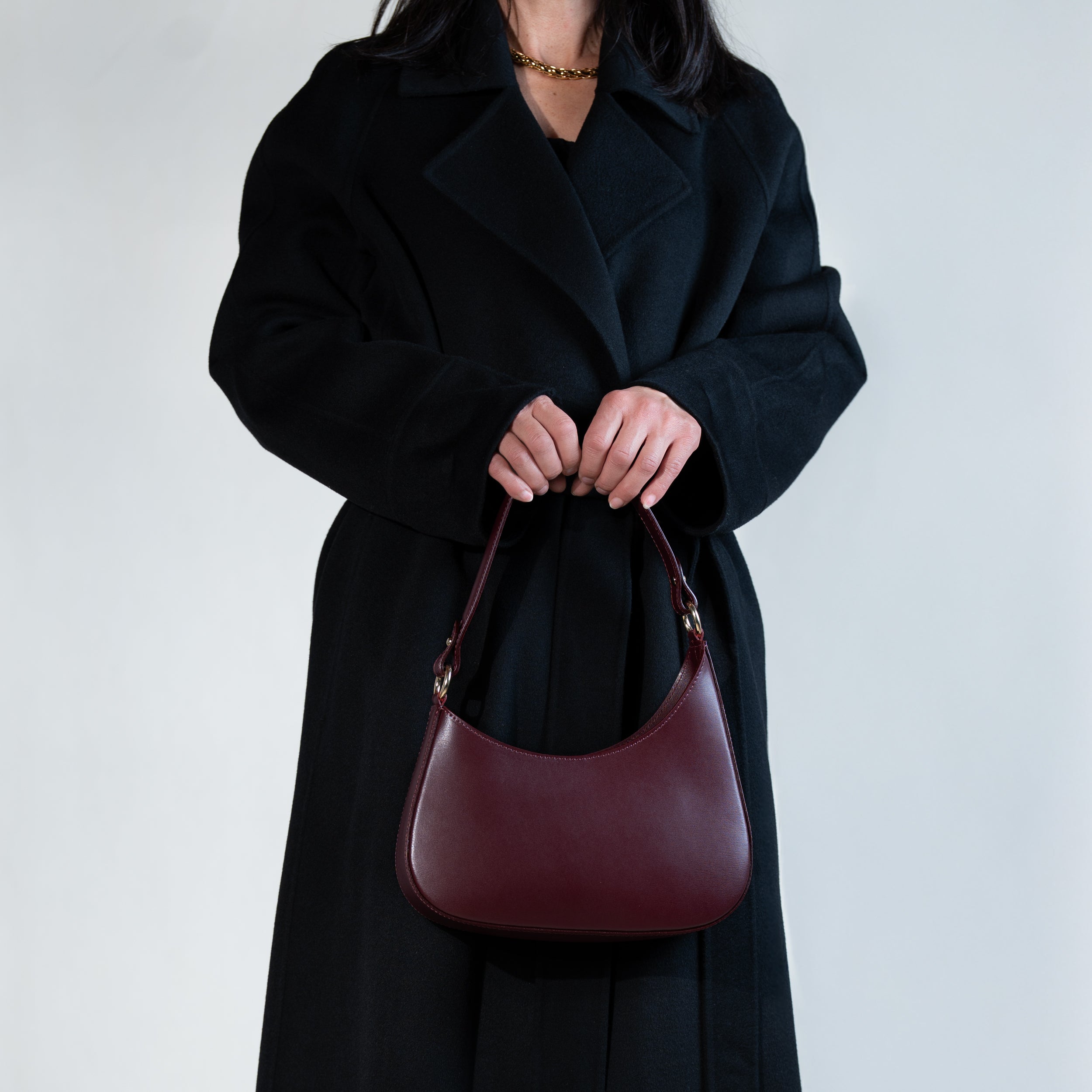 sac à main forme hobo demi lune asymétrique pour femme en cuir lisse bordeaux porté à la main de dos sur manteau noir 