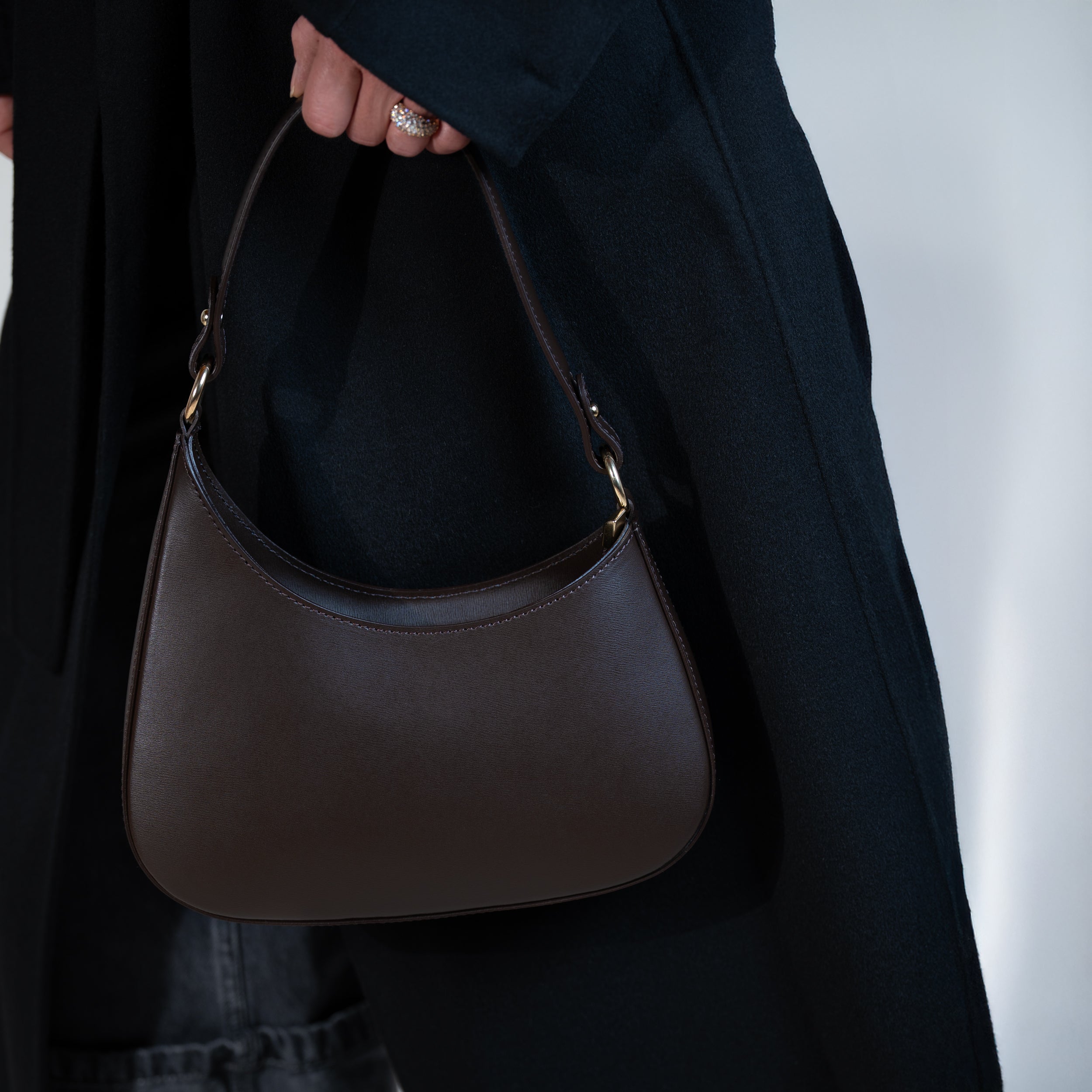 sac à main forme hobo demi lune asymétrique pour femme en cuir lisse marron porté à la main sur un manteau noir