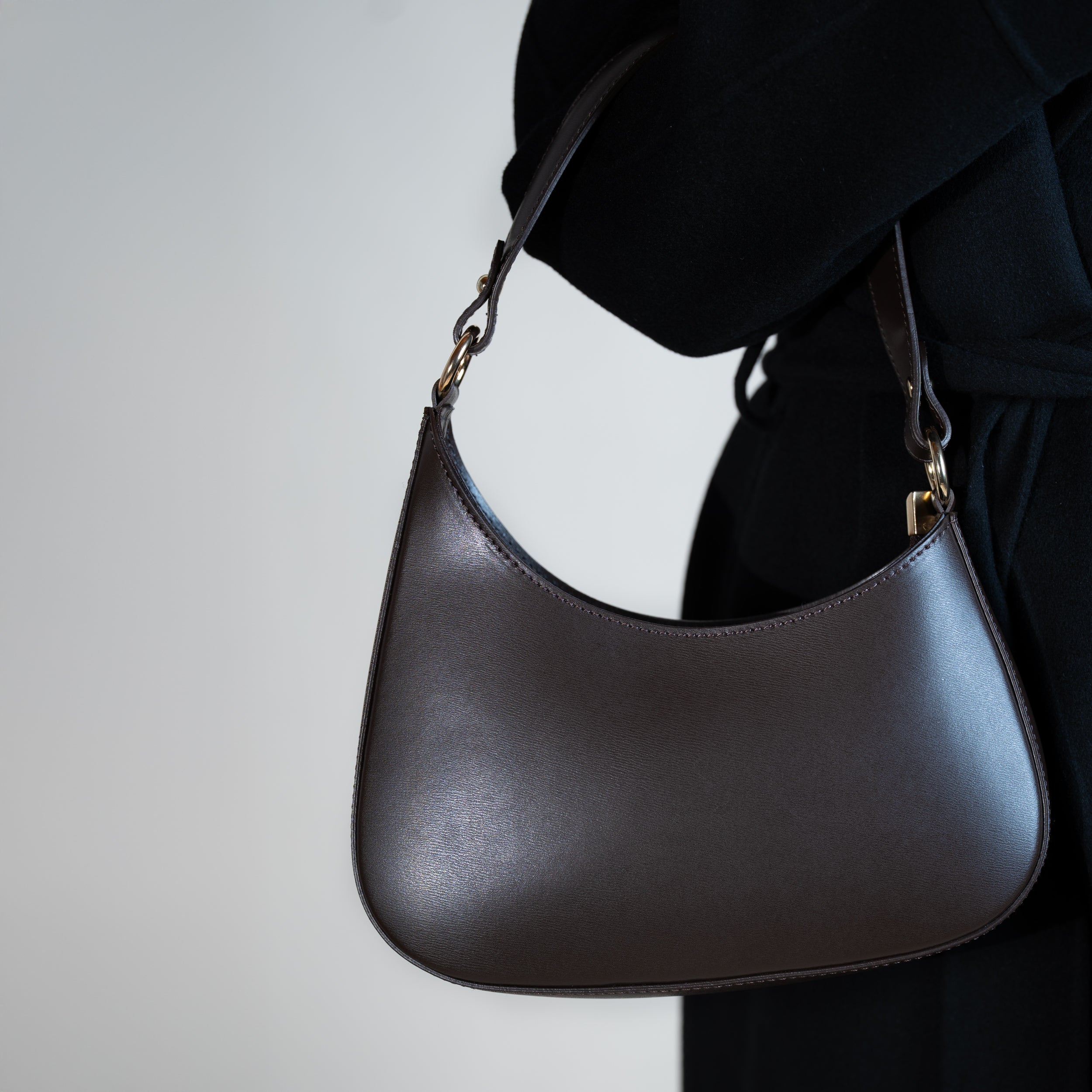 sac à main forme hobo demi lune asymétrique pour femme en cuir lisse marron porté au creux de bras sur un manteau noir