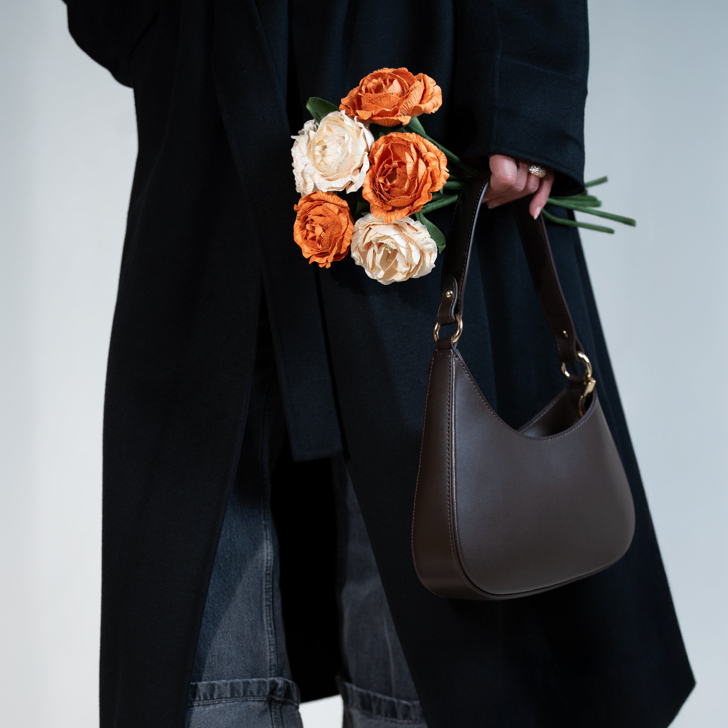 sac à main forme hobo demi lune asymétrique pour femme en cuir lisse marron porté à la main sur un manteau noir avec bouquet de camélia rose et orange