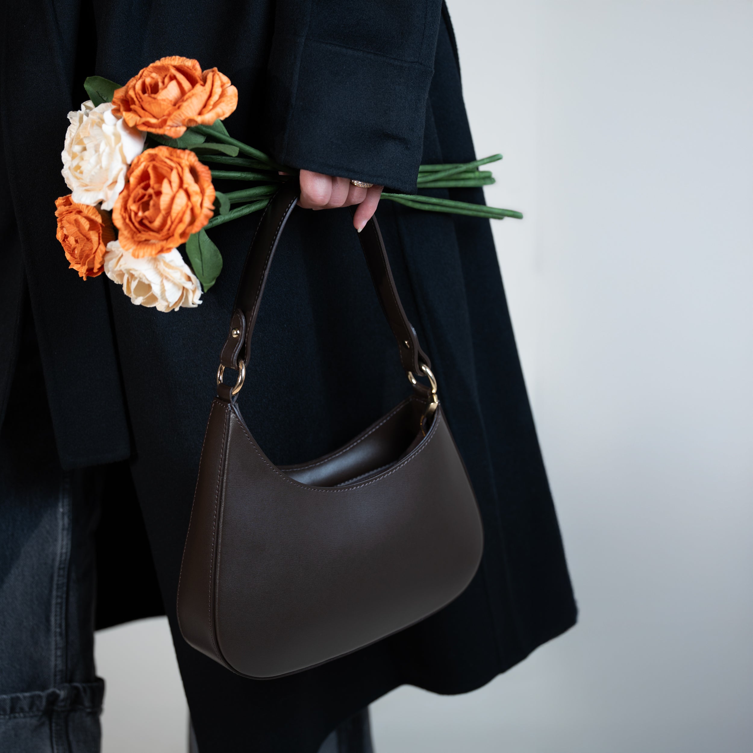 sac à main forme hobo demi lune asymétrique pour femme en cuir lisse marron porté à la main sur un manteau noir et jean noir