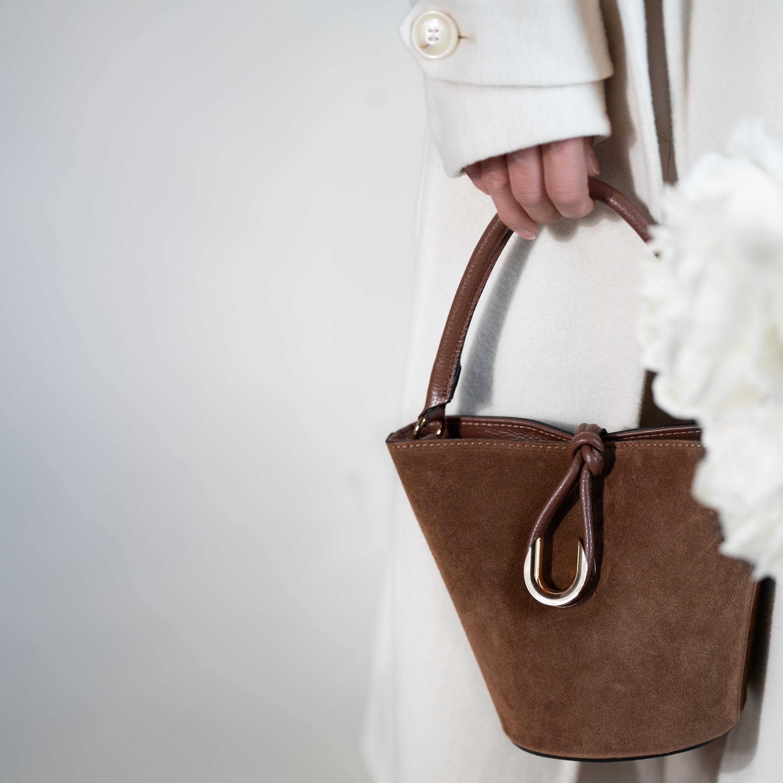 sac à main femme en cuir suédé ou daim brun en forme seau avec un fermoir doré original porté à la main sur un manteau blanc avec des pivoine blanche