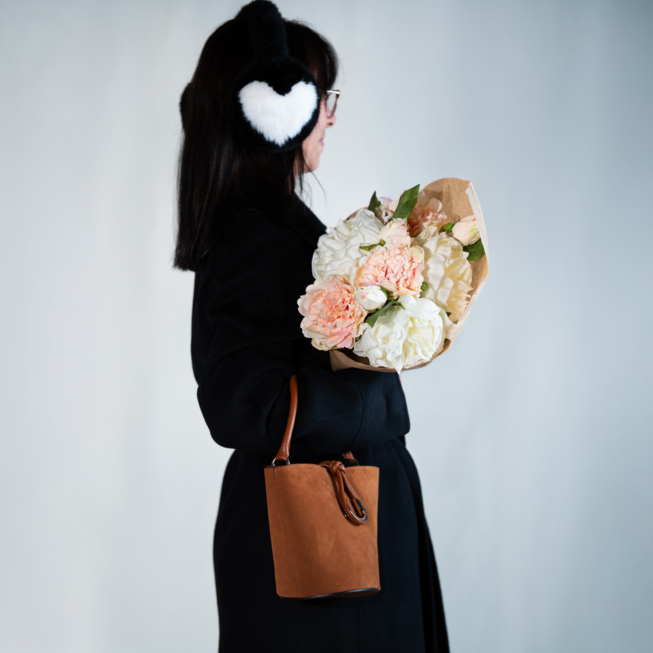 sac à main femme en cuir suédé ou daim camel en forme seau porté au creux de bras avec un fermoir doré original sur un manteau noir avec un bouquet de pivoine et un cache oreille coeur