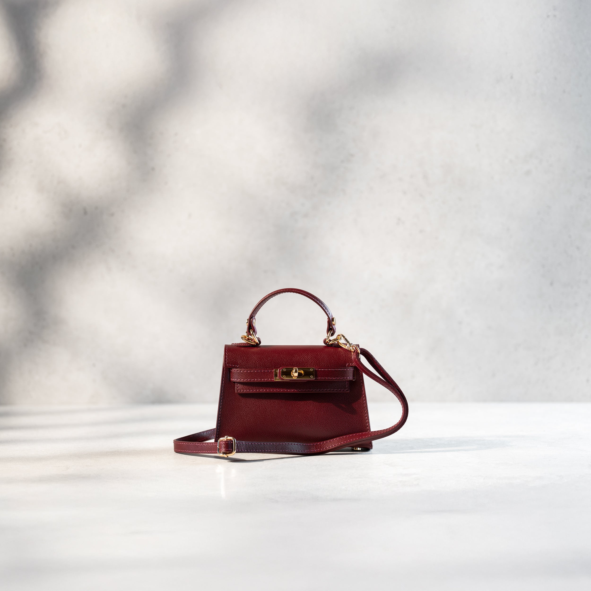 sac à main femme en cuir saffiano bordeaux top handle avec une bandoulière, un fermoir tourniquet, une bijouterie doré et des clous de fond de sac 