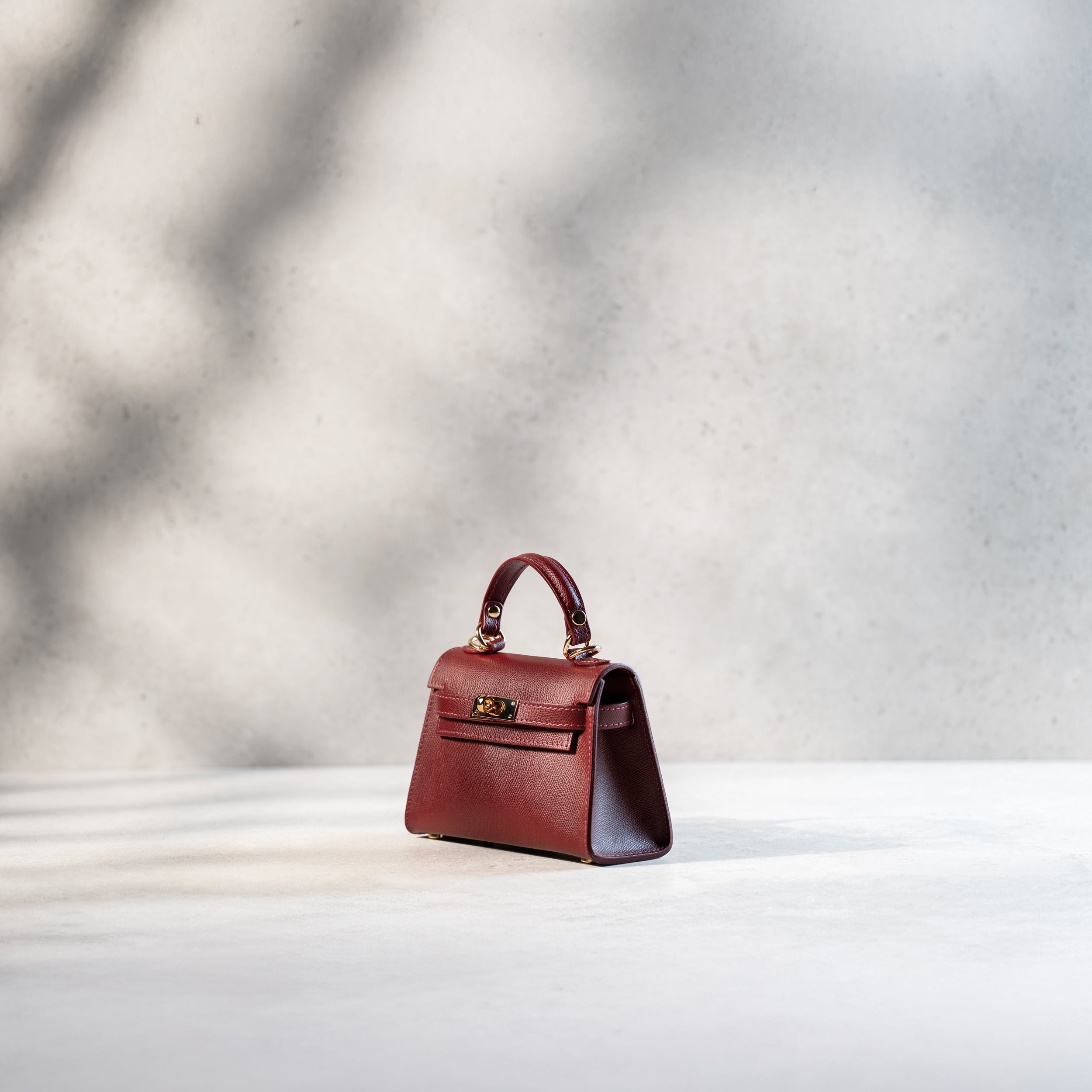 sac à main femme en cuir saffiano bordeaux top handle de côté avec un fermoir tourniquet, une bijouterie doré et des clous de fond de sac 