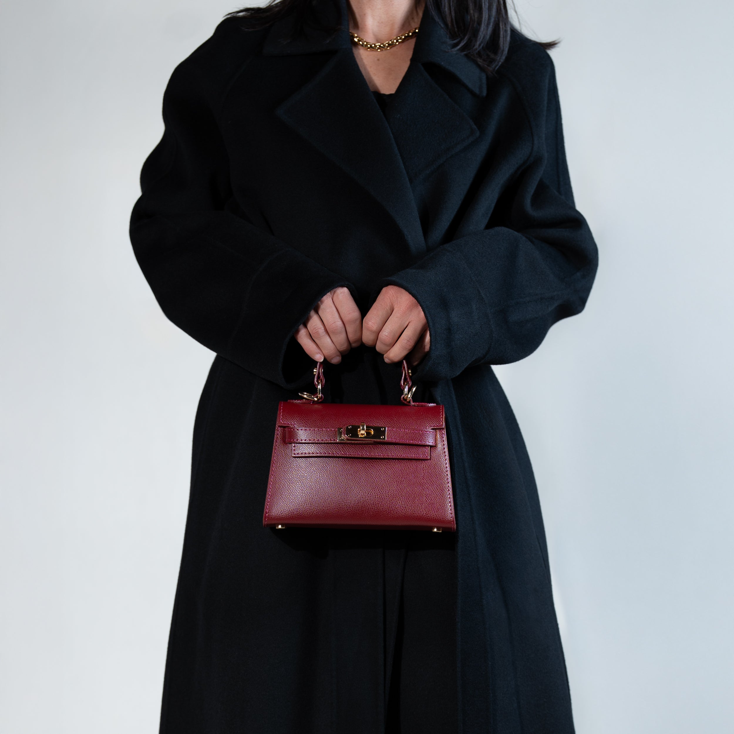 sac à main femme en cuir saffiano bordeau et bijouterie dorée dans une forme top handle porté à la main avec un manteau noir 