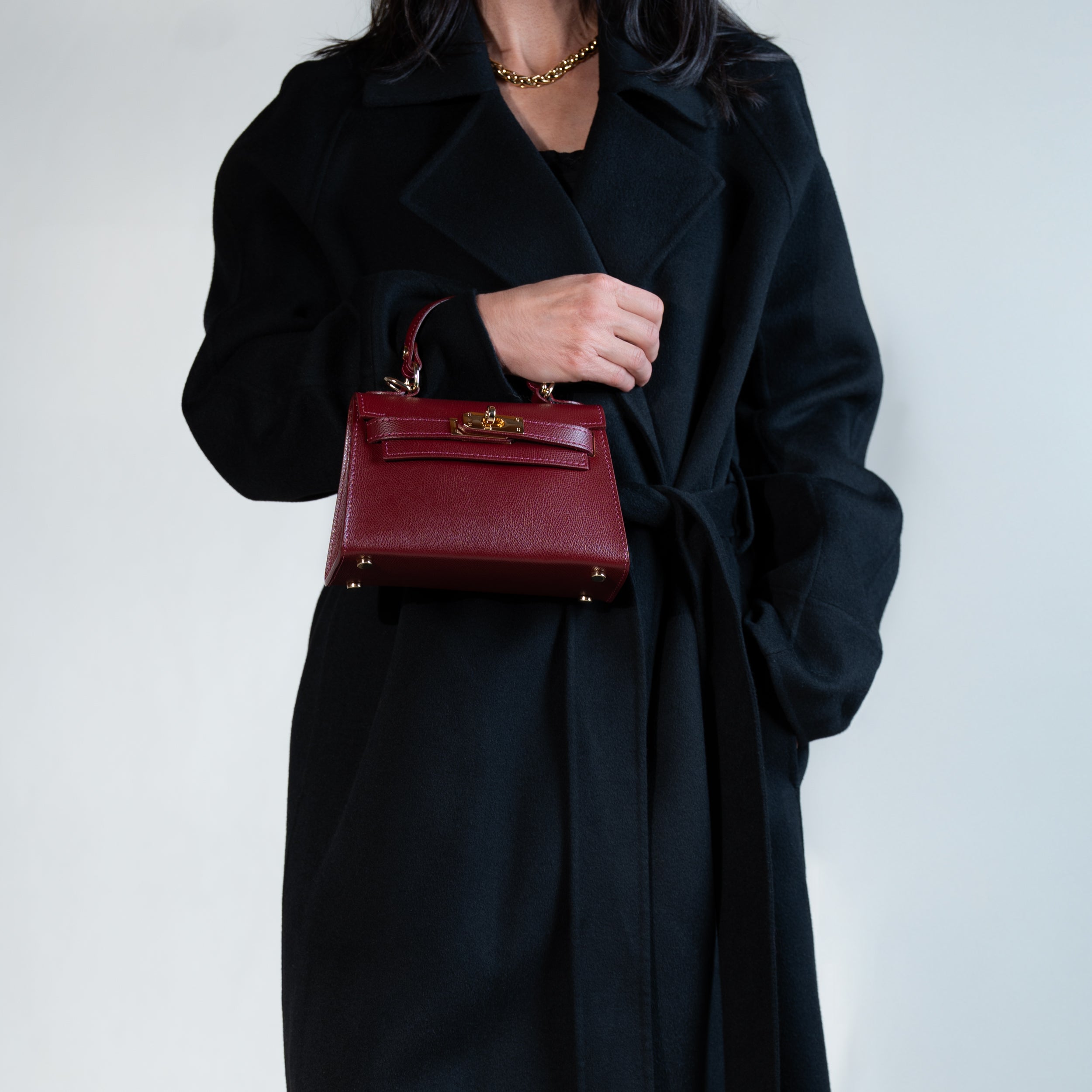 sac à main femme en cuir saffiano top handle porté au bras sur un manteau noir  avec fermoir tourniquet et bijouterie doré dans un cuir de qualité bordeaux et clous de fond de sac 