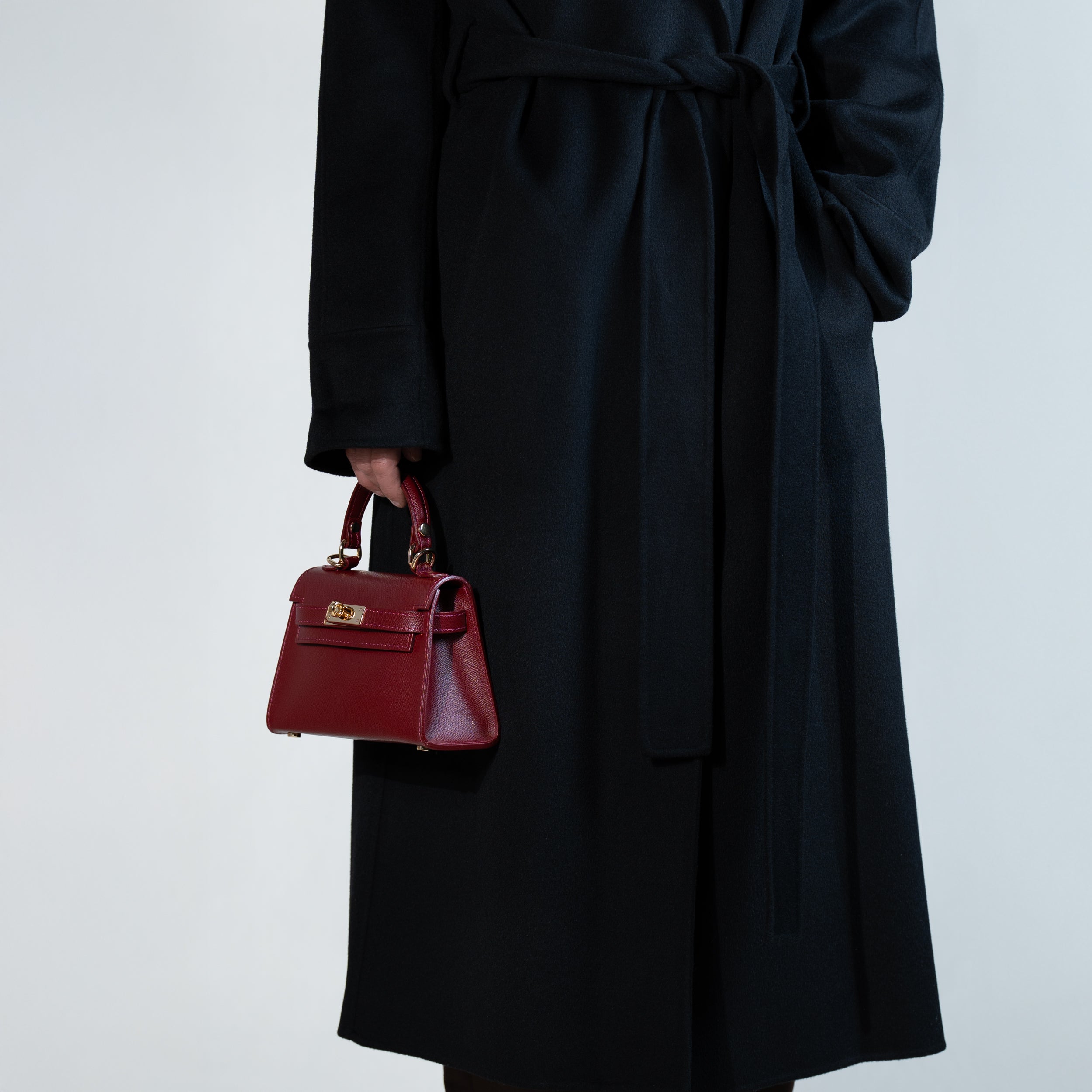 sac à main femme en cuir saffiano top handle porté à la main sur un manteau noir  avec fermoir tourniquet et bijouterie doré dans un cuir de qualité bordeaux 