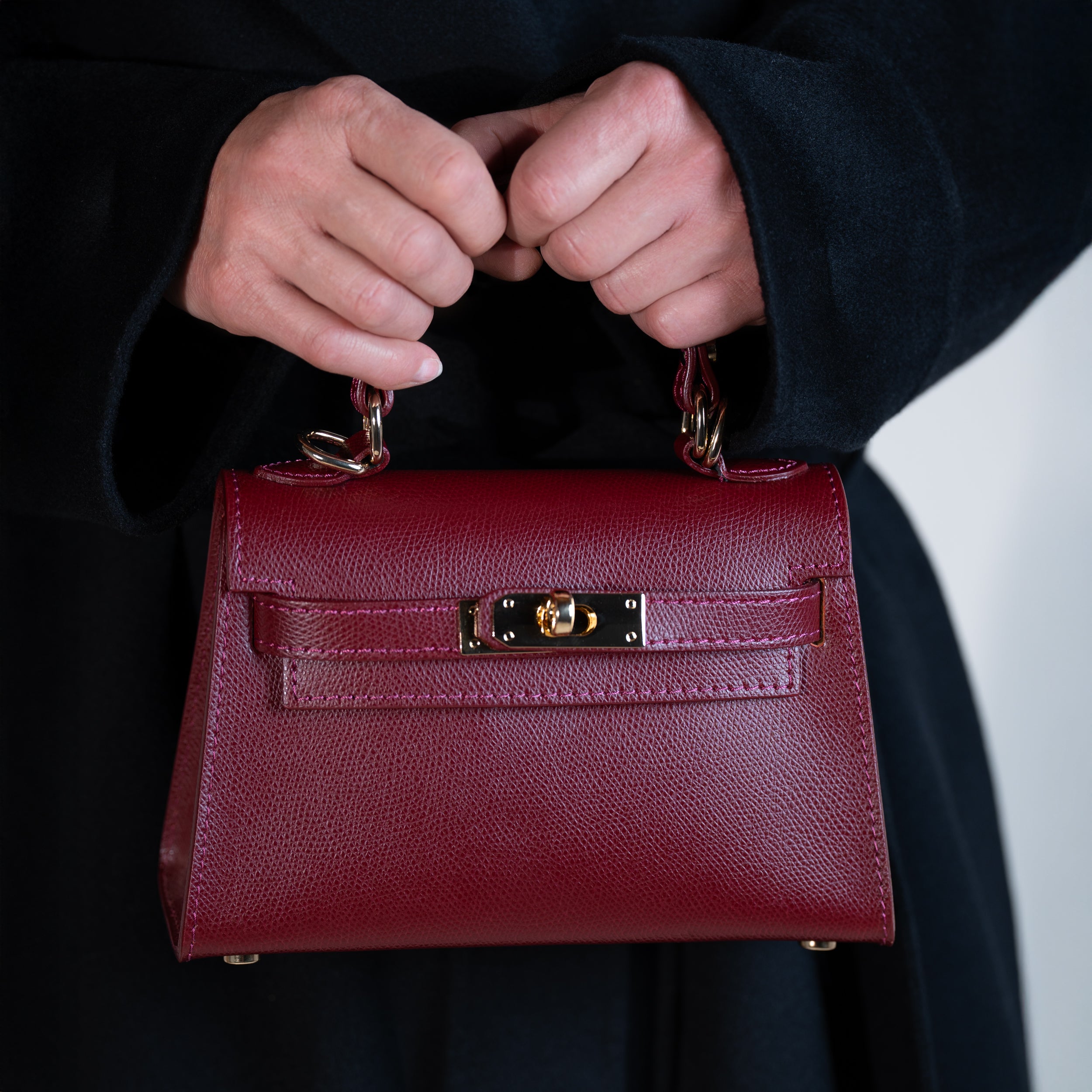 sac à main femme en cuir saffiano top handle avec fermoir tourniquet et bijouterie doré dans un cuir de qualité bordeaux porté à la main sur un manteau noir 
